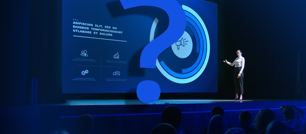 A woman standing on stage with a giant question mark in front of an event presentation.